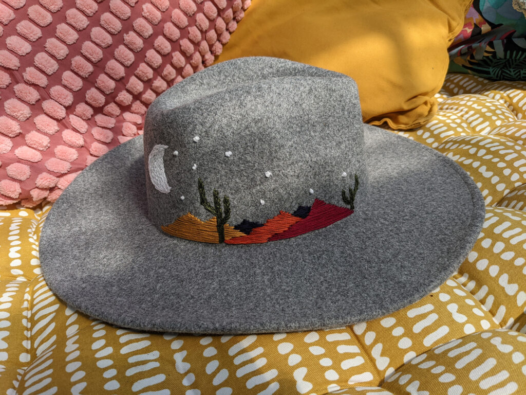 Image shows grey cowboy hat sitting on a yellow cushion. On the hat a landscape feating mountains and cacti have been hand embroidered. A crescent moon sits on the left hand side above the mountain. Stars are in the sky. 