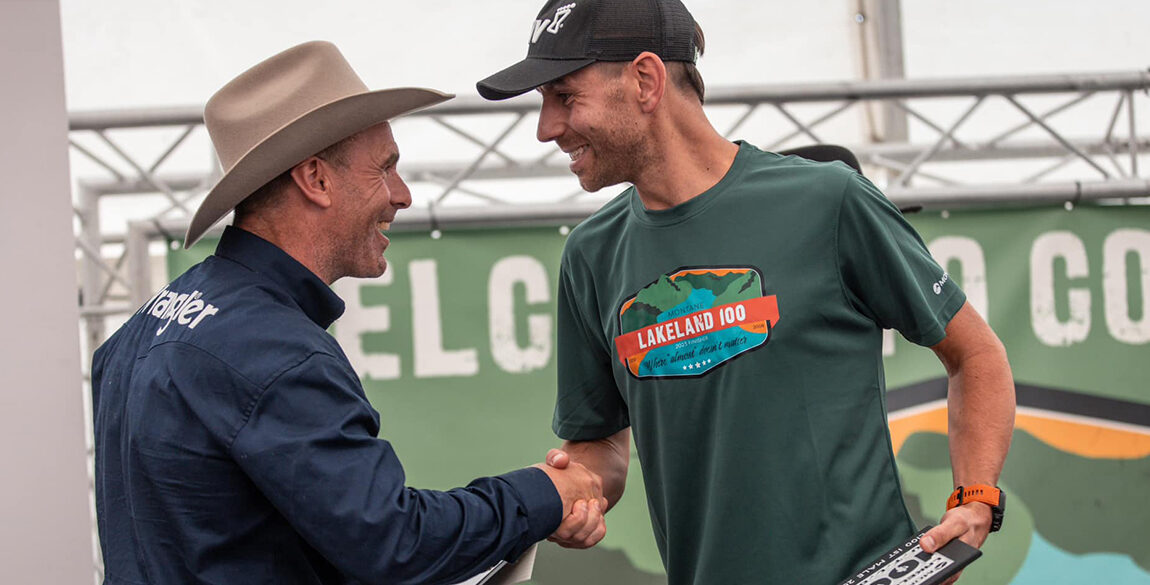 Image shows the event organiser shaking the hand of the male 100 winner on stage. He wears the 2023 green finishers tshirt.