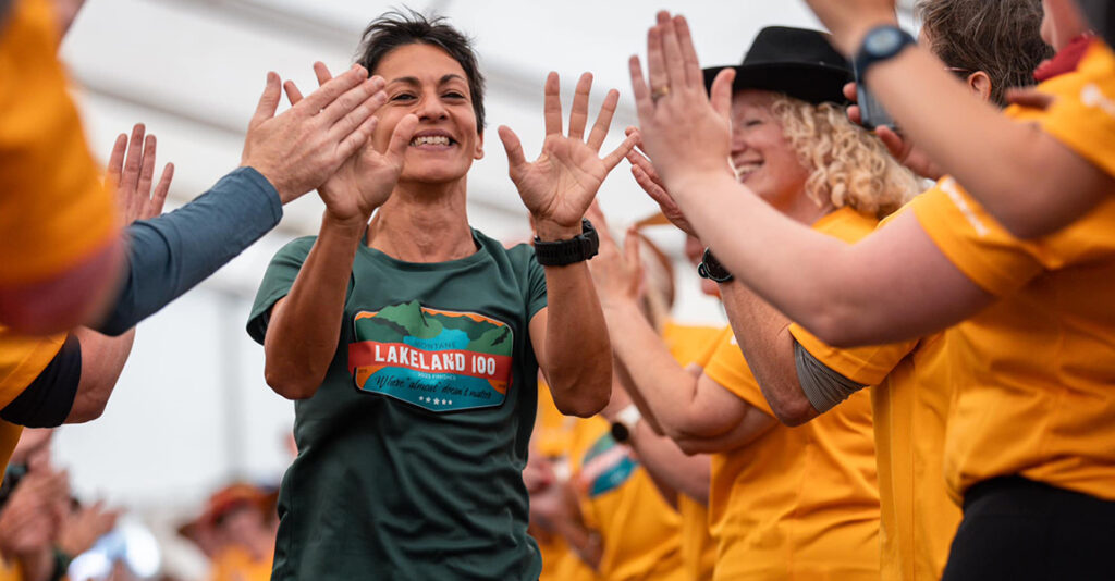 Images shows the Lakeland 2023 100 female winner coming through a crowd of volunteers. She wears the green finishers tshirt with Lakeland 2023 logo.