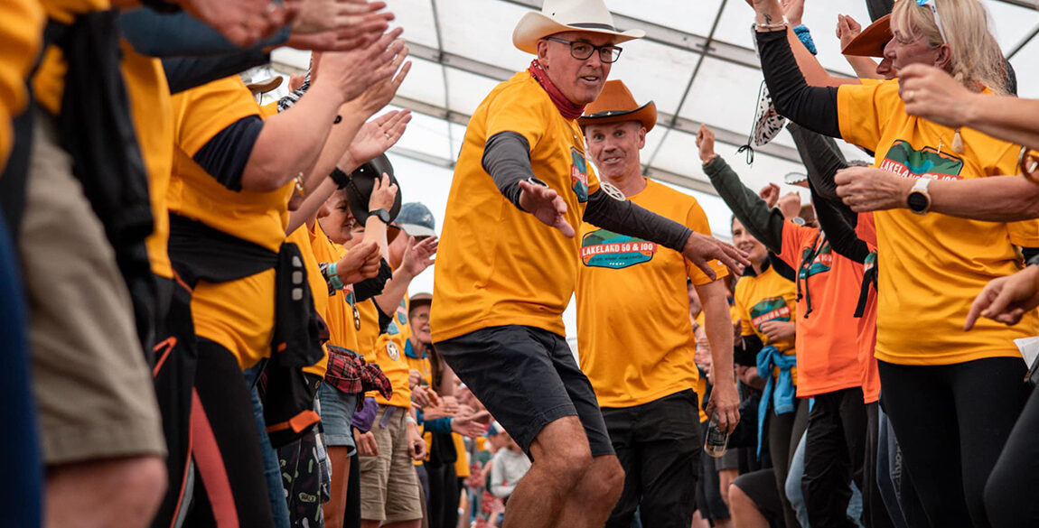Image shows one of the volunteers dancing through a crowd of other volunteers, all wearing their yellow tshirts with Lakeland 2023 event logo on
