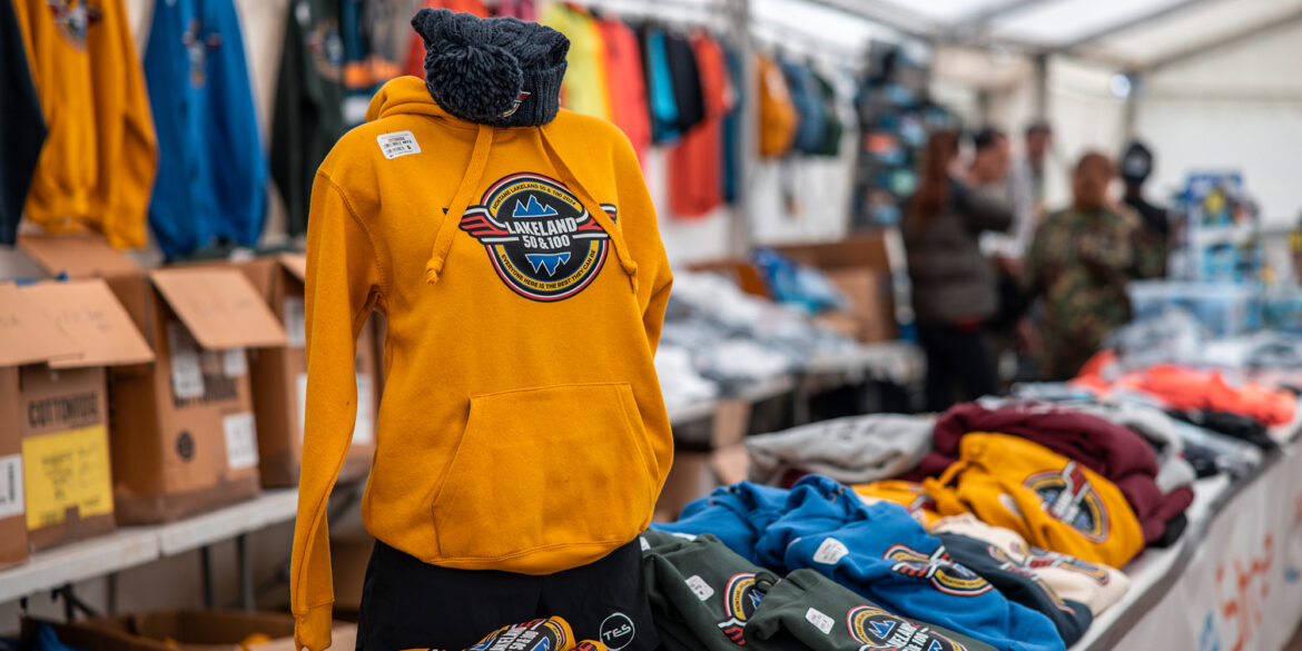 A merchandise stand for the Montane Lakeland 50 and 100 event. A yellow hoodie with the event logo is prominently displayed, with a black beanie on top. In the foreground and background, various colored shirts and hoodies with the same logo are visible. The stand is inside a white tent, with cardboard boxes and folded clothing on shelves and tables.