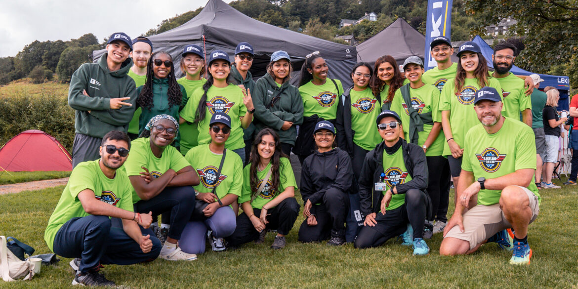 A group photo of diverse volunteers at the Lakeland 50 and 100 event. About 20 people are posing together, smiling and wearing bright green t-shirts with the event logo on. They're standing in front of tents on a grassy field with trees and hills in the background. Some wear caps and sunglasses.