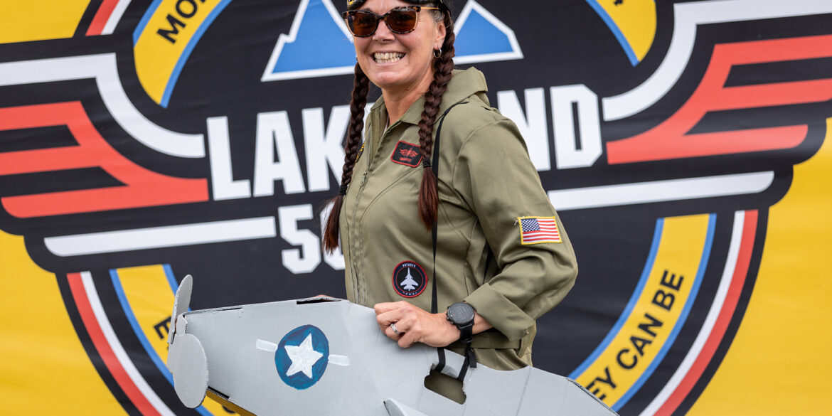 A smiling woman with braided hair stands in front of a large Montane Lakeland 50 & 100 2024 logo. She's wearing an olive green flight suit with patches, including an American flag, and has sunglasses on her head. She's holding a cardboard cutout of a fighter jet.