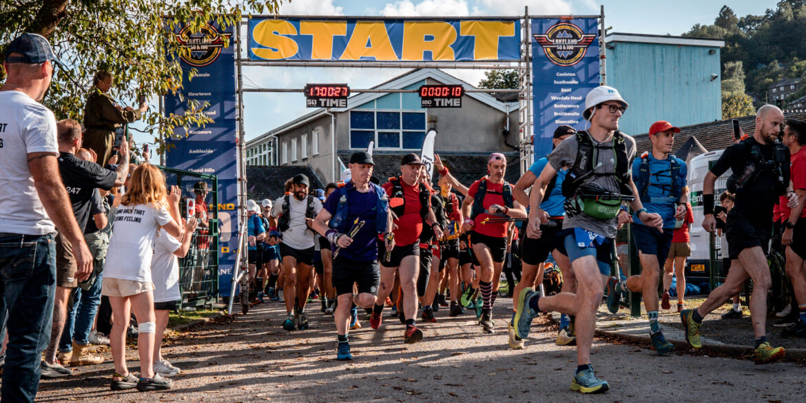 The start of the Montane Lakeland 100 race. Runners are passing under a large 'START' banner with digital timers showing 17:00:37 for the 50 mile race and 0:00:10 for the 100 mile race. Participants wearing colorful running gear and backpacks are beginning their journey. Spectators line the sides, cheering them on. The scene is set on a sunny day with trees and buildings visible in the background, capturing the excitement and energy of the race's beginning.