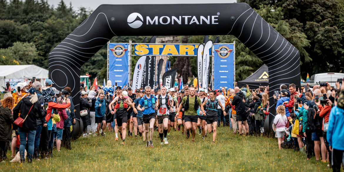 The start of the Montane Lakeland 50 race. Runners are passing under a large black inflatable arch branded with 'MONTANE'. Behind it is a 'START' banner. A group of runners in athletic gear with backpacks are beginning their race, surrounded by spectators on both sides. The scene is set in a grassy field with trees in the background, capturing the excitement of the race's beginning.