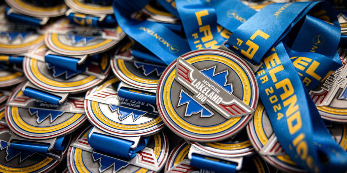 Close-up of numerous silver medals for the 'Montane Lakeland 100' event. The medals feature a blue mountain logo and are attached to bright blue ribbons with yellow text reading 'LAKELAND 100'. Many medals are stacked together, with one prominently displayed on top showing the full design.