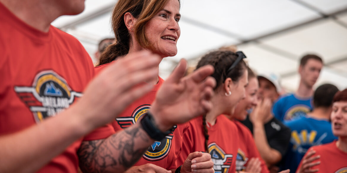 A group of people in red shirts with the Lakeland 100 and 50 event logo on, standing in a tent. The focus is on a smiling woman with brown hair in the center. Beside her, a person's tattooed arm is visible. The group appears to be clapping or cheering. In the background, some people are wearing blue shirts with the same logo.