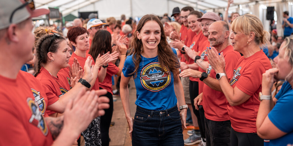 A Montane Lakeland 50 finisher, a smiling woman with long brown hair wearing a blue event t-shirt, stands in the center of a crowd. She is surrounded by event volunteers in red shirts with the same logo, who are applauding and cheering. The scene takes place inside a large tent, at the event's finisher ceremony. The atmosphere is joyful and congratulatory.