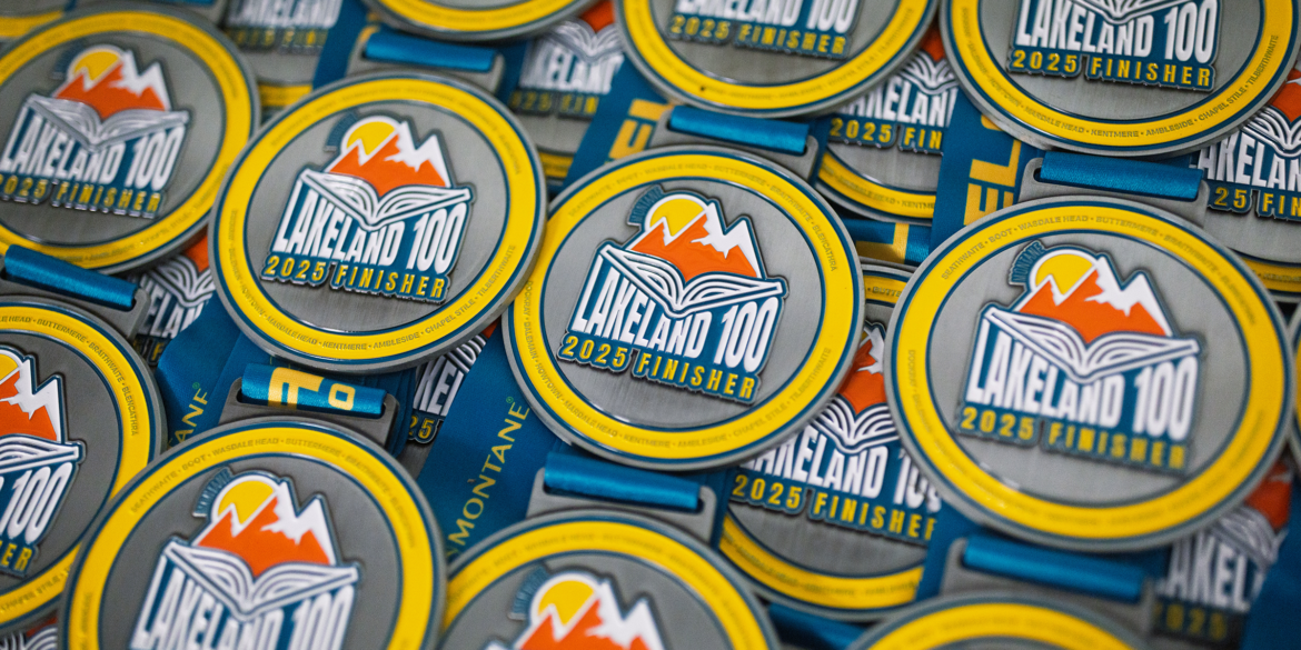 Close-up of Lakeland 100 finisher medals featuring the Lakeland 2025 event logo design with mountain and storybook motif in orange, yellow, and teal.