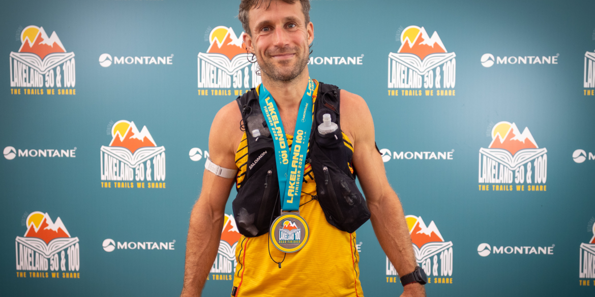 Lakeland 100 male finisher wearing medal featuring the Lakeland 2025 event logo design, standing in front of branded backdrop with event graphics.
