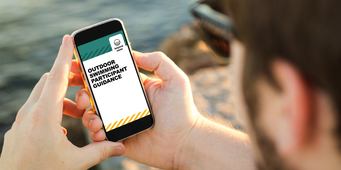 A swimmer holding a phone displaying the Beyond Swim Outdoor Swimming Participant Guidance, designed as part of open-water swimming venue design guidance.